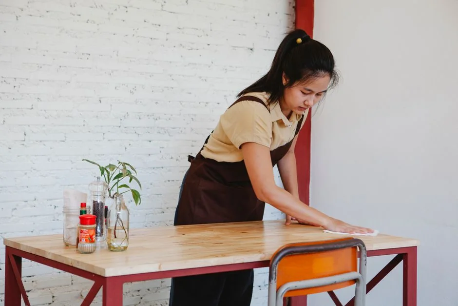 A woman wearing a plaid shirt cleans a kitchen counter using a spray bottle and cloth.